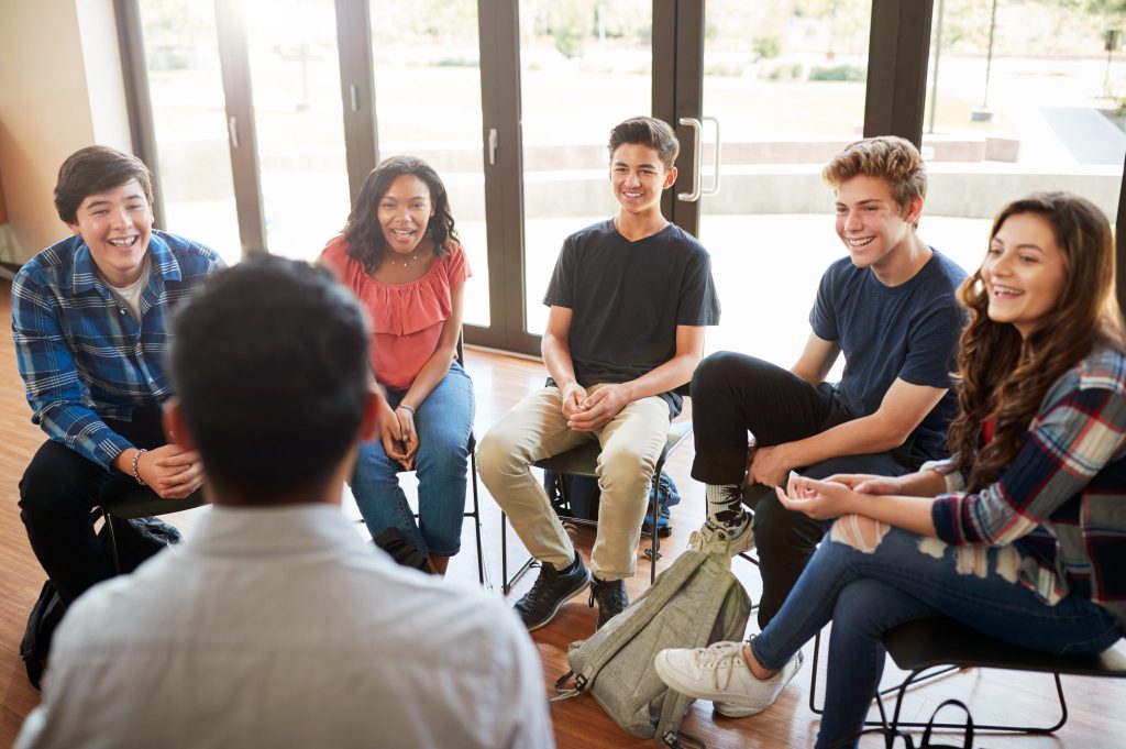 A group of young people sitting in a semi circle around an adult, smiling and laughing.